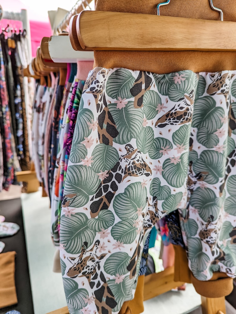 May include: Pants featuring a white background with a pattern of green tropical leaves, small pink flowers, and brown giraffe heads. The garment has a solid brown elastic waistband and matching brown elastic cuffs at the bottom of the legs.