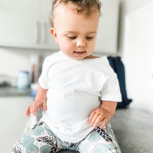 May include: A plain white short-sleeved t-shirt paired with leggings featuring a repeating pattern of giraffe heads and light green tropical leaves on a white background. The leggings have a fitted silhouette and a colorful animal print design.