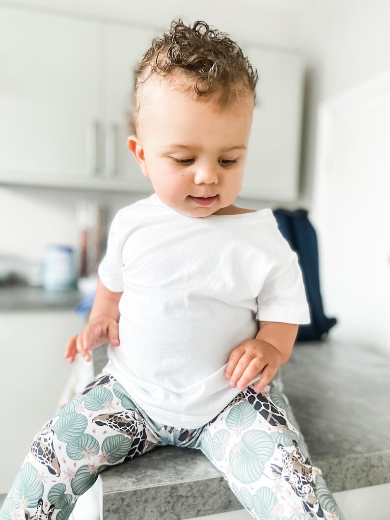 May include: A plain white short-sleeved t-shirt paired with leggings featuring a repeating pattern of giraffe heads and light green tropical leaves on a white background. The leggings have a fitted silhouette and a colorful animal print design.