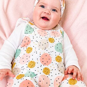 K&ouml;nnte beinhalten: Ein wei&szlig;er Langarm-Strampler mit einem Muster aus rosa, mintgr&uuml;nen und goldglitzernden Kreisen sowie kleinen schwarzen Punkten. Der Strampler wird mit einem passenden geknoteten Stirnband kombiniert, das dasselbe Kreis- und Punktemuster aufweist.