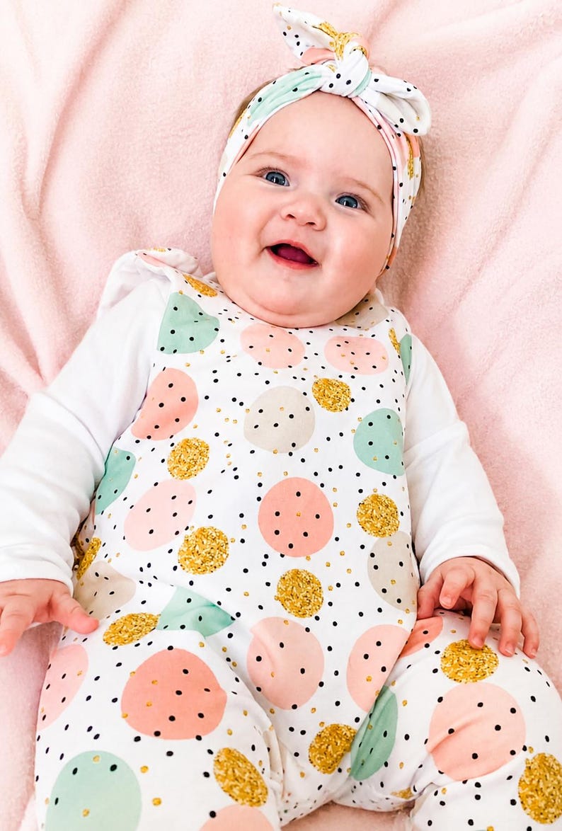 K&ouml;nnte beinhalten: Ein wei&szlig;er Langarm-Strampler mit einem Muster aus rosa, mintgr&uuml;nen und goldglitzernden Kreisen sowie kleinen schwarzen Punkten. Der Strampler wird mit einem passenden geknoteten Stirnband kombiniert, das dasselbe Kreis- und Punktemuster aufweist.