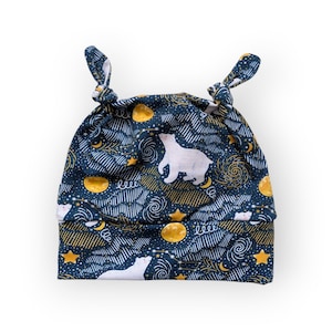 May include: A navy blue baby hat with a whimsical night sky pattern. The design includes white polar bears, golden moons, stars, and swirling cloud-like shapes. The hat has two knotted ties at the top.