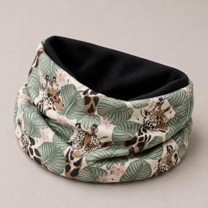May include: A neck warmer featuring a cream-colored exterior with a repeating pattern of brown and tan giraffe heads, green tropical leaves, and small pink flowers. The interior of the circular accessory is lined with solid black fabric.