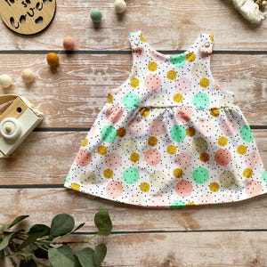 K&ouml;nnte beinhalten: Ein &auml;rmelloses Babykleid mit wei&szlig;em Hintergrund und einem Muster aus verstreuten Punkten in Minzgr&uuml;n, zartem Rosa, Creme und glitzerndem Gold. Das Kleid hat eine geraffte Taille und zwei metallische Druckknopfverschl&uuml;sse an den Schultern.