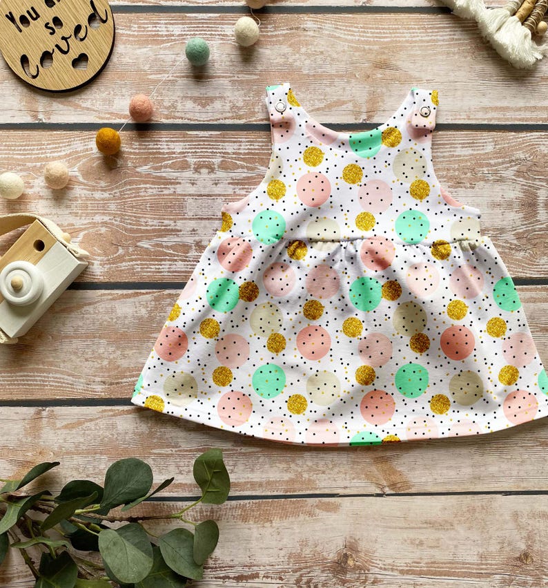 K&ouml;nnte beinhalten: Ein &auml;rmelloses Babykleid mit wei&szlig;em Hintergrund und einem Muster aus verstreuten Punkten in Minzgr&uuml;n, zartem Rosa, Creme und glitzerndem Gold. Das Kleid hat eine geraffte Taille und zwei metallische Druckknopfverschl&uuml;sse an den Schultern.