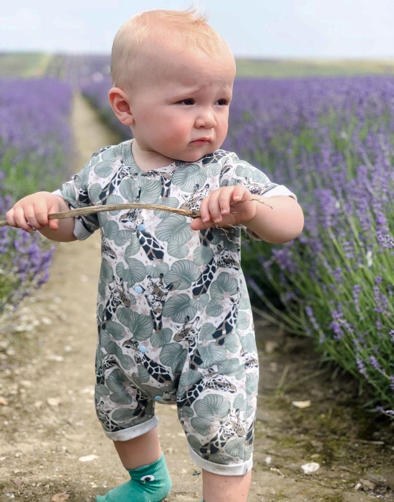 May include: A short-sleeved romper featuring a pattern of giraffes and green tropical leaves. The garment has a round neckline, light blue snap buttons down the front, and white trim on the sleeves and leg openings. The fabric has a light background.