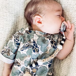 May include: A baby wearing a short-sleeved romper with a green and white jungle-themed print featuring giraffes and leaves. A pacifier clip with black, white, and wooden beads spells out the name FORREST and attaches to the romper with a metal clip.