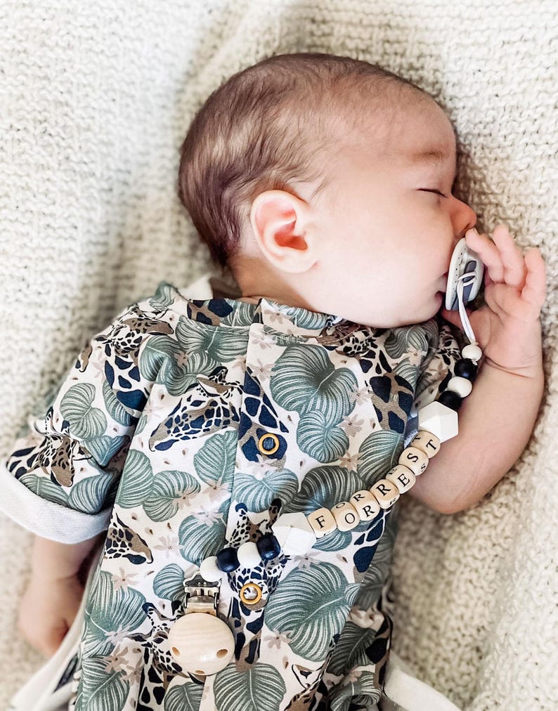 May include: A baby wearing a short-sleeved romper with a green and white jungle-themed print featuring giraffes and leaves. A pacifier clip with black, white, and wooden beads spells out the name FORREST and attaches to the romper with a metal clip.