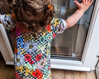 May include: A young child wearing a colourful floral and chequered jumpsuit with a black and white chequered pattern. The child is standing near a window and reaching out with their hand.