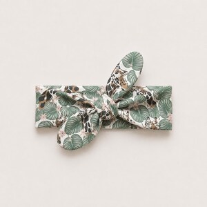 May include: A fabric headband featuring a knotted bow detail at the center. The material is patterned with a repeating design of green tropical leaves, small pink flowers, and brown and white giraffe print spots against a white background.