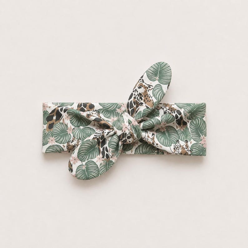 May include: A fabric headband featuring a knotted bow detail at the center. The material is patterned with a repeating design of green tropical leaves, small pink flowers, and brown and white giraffe print spots against a white background.
