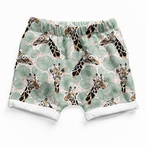 May include: Shorts featuring an all-over print of giraffe heads and light green tropical leaves on a pale background. The garment has an elasticized waistband and white cuffed leg openings, providing a structured look with a repeating pattern.