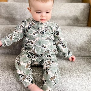 May include: A long-sleeved button-down shirt and matching pants set featuring a repeating pattern of giraffes and large green tropical leaves on a white background. The garment has a round neckline and a front button closure with small white buttons.
