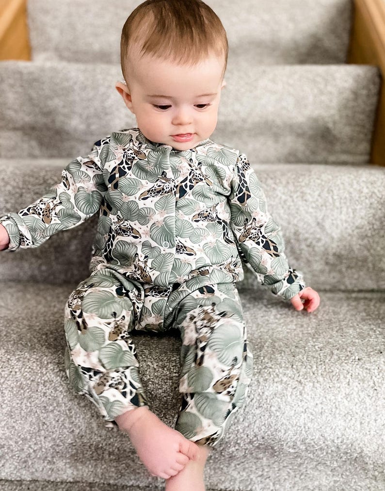 May include: A long-sleeved button-down shirt and matching pants set featuring a repeating pattern of giraffes and large green tropical leaves on a white background. The garment has a round neckline and a front button closure with small white buttons.