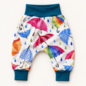 May include: Pull-on trousers featuring a white background with a colourful watercolour print of various umbrellas and raindrops. The garment includes a wide, solid dark teal elastic waistband and matching dark teal elastic cuffs at the ankles.