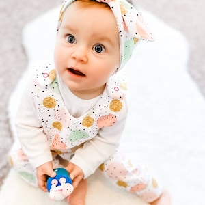 K&ouml;nnte beinhalten: Ein wei&szlig;es Langarmshirt und eine passende Latzhose mit einem Muster aus goldglitzernden, rosafarbenen und mintgr&uuml;nen Kreisen mit schwarzen Punkten. Das Outfit enth&auml;lt ein passendes geknotetes Stirnband mit demselben Kreis- und Punktemuster.