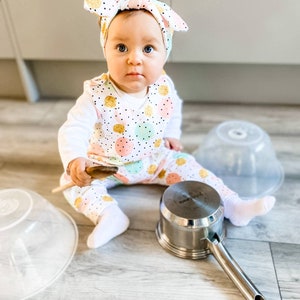 K&ouml;nnte beinhalten: Ein wei&szlig;er Langarm-Strampler mit einem Muster aus rosa, t&uuml;rkisfarbenen und goldglitzernden Kreisen mit schwarzen Punkten, kombiniert mit einem passenden geknoteten Stirnband. Das Outfit wird mit wei&szlig;en Socken und einem Holzl&ouml;ffel getragen.
