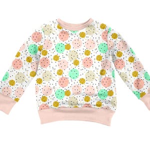 K&ouml;nnte beinhalten: Ein lang&auml;rmeliges Sweatshirt mit wei&szlig;em Hintergrund und einem wiederkehrenden Muster aus hellrosa, mintgr&uuml;nen und goldglitzernden Kreisen. Das Design enth&auml;lt kleine schwarze Sprenkel und ist mit einem einfarbigen hellrosa Rundhalsausschnitt, B&uuml;ndchen und Saum versehen.