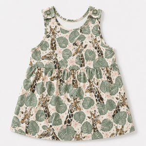 May include: A sleeveless dress featuring a repeating pattern of giraffes, green tropical leaves, and small pink flowers on a cream background. The garment has a gathered waistline and two metallic snap buttons on the shoulder straps for closure.