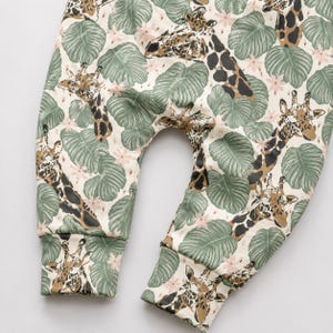 May include: Pants featuring a cream background with a repeating pattern of brown and tan giraffes interspersed with sage green tropical monstera leaves and small pale pink flowers. The garment has cuffed ankles and a curved crotch seam.