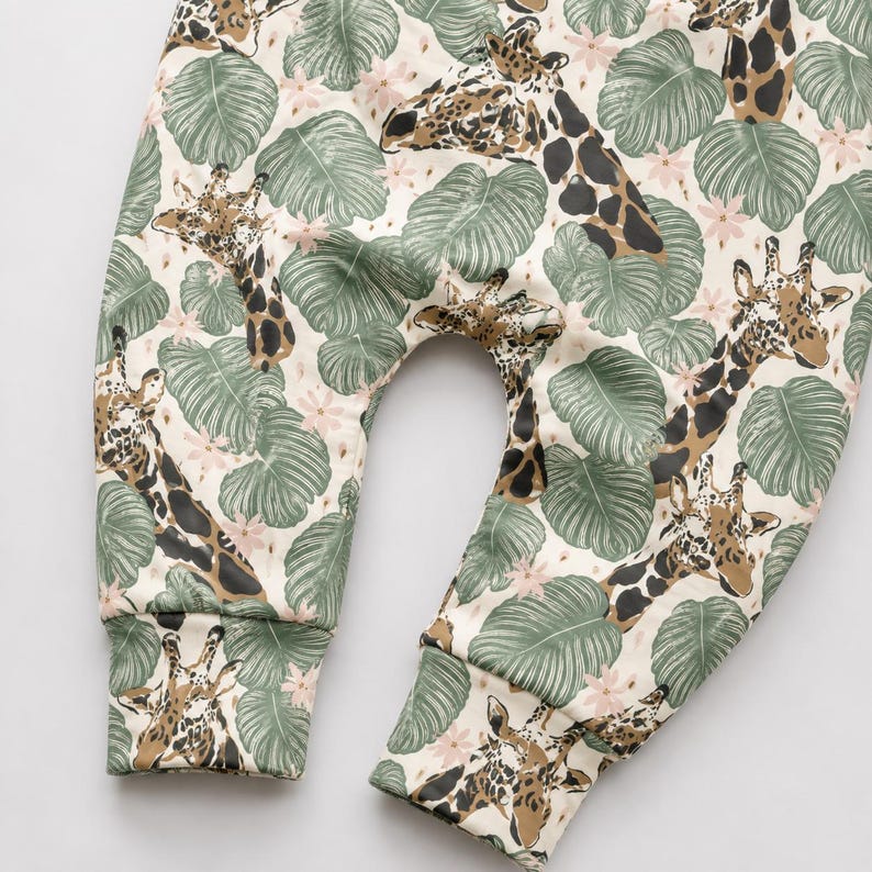 May include: Pants featuring a cream background with a repeating pattern of brown and tan giraffes interspersed with sage green tropical monstera leaves and small pale pink flowers. The garment has cuffed ankles and a curved crotch seam.