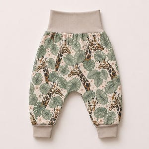 May include: Pants featuring a repeating pattern of brown and tan giraffes interspersed with sage green tropical leaves on a cream background. The garment includes a wide, solid beige elastic waistband and matching beige ribbed ankle cuffs.