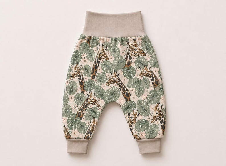 May include: Pants featuring a repeating pattern of brown and tan giraffes interspersed with sage green tropical leaves on a cream background. The garment includes a wide, solid beige elastic waistband and matching beige ribbed ankle cuffs.