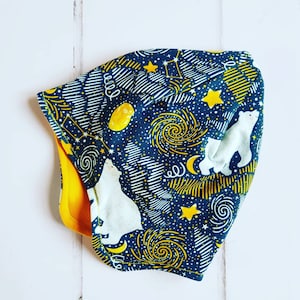 May include: A blue and yellow fabric hat with a polar bear design. The hat has a yellow lining and a swirling pattern of stars and moons.