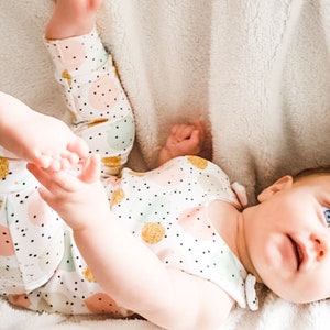 K&ouml;nnte beinhalten: Ein Baby-Outfit bestehend aus einem &auml;rmellosen Oberteil und einer Hose mit wei&szlig;em Hintergrund, bedruckt mit kleinen schwarzen Punkten und gr&ouml;&szlig;eren Kreisen in Pastellrosa, Mintgr&uuml;n und Goldglitzer. Die Kleidung wird von einem Baby mit blauen Augen getragen.