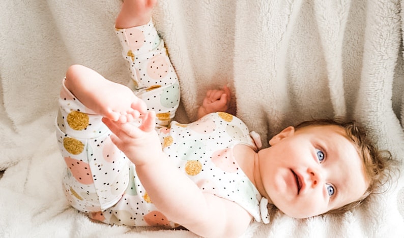 K&ouml;nnte beinhalten: Ein Baby-Outfit bestehend aus einem &auml;rmellosen Oberteil und einer Hose mit wei&szlig;em Hintergrund, bedruckt mit kleinen schwarzen Punkten und gr&ouml;&szlig;eren Kreisen in Pastellrosa, Mintgr&uuml;n und Goldglitzer. Die Kleidung wird von einem Baby mit blauen Augen getragen.