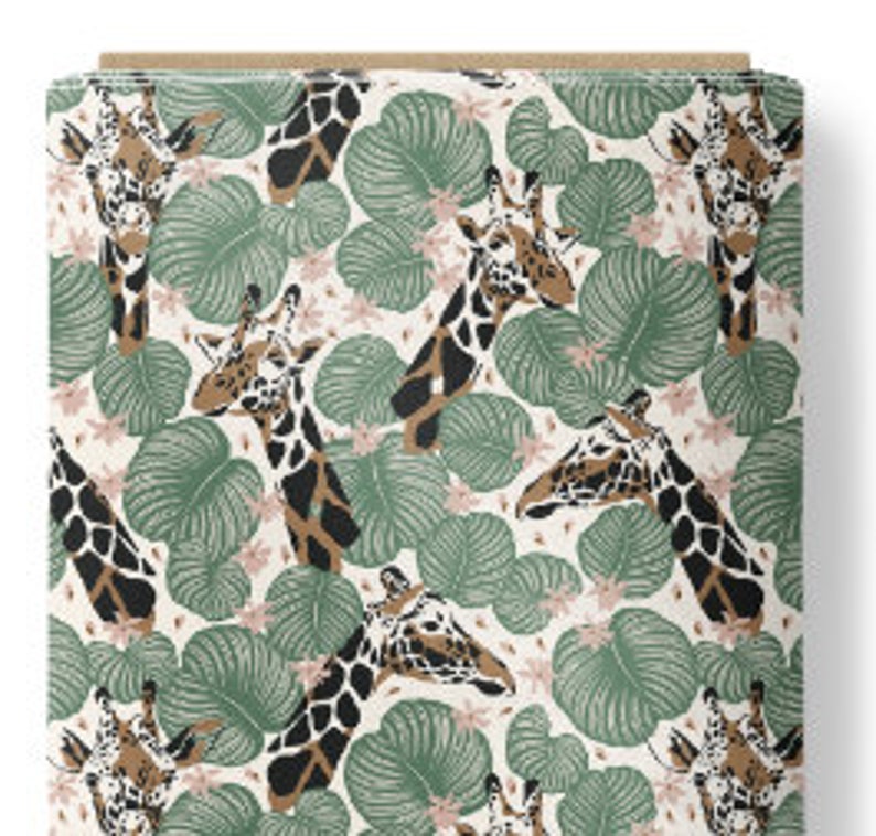 May include: A bolt of fabric featuring a repeating pattern of giraffe heads interspersed with large green tropical leaves and small light pink flowers on a cream-colored background. The giraffes have brown and black spots and white faces.