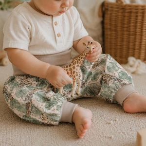 May include: A short-sleeved cream-colored ribbed shirt with three buttons at the neckline and pants featuring a pattern of green tropical leaves and giraffes. The pants have wide, solid light brown elastic cuffs at the waist and ankles.