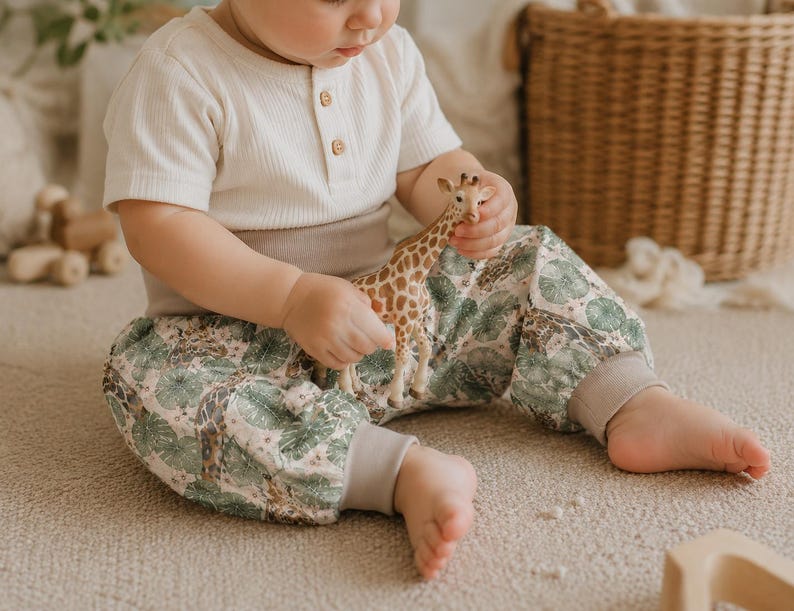 May include: A short-sleeved cream-colored ribbed shirt with three buttons at the neckline and pants featuring a pattern of green tropical leaves and giraffes. The pants have wide, solid light brown elastic cuffs at the waist and ankles.