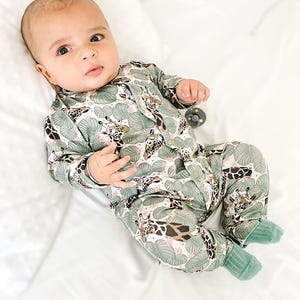 May include: A long-sleeved baby romper featuring a repeating pattern of giraffes and large green tropical leaves on a light background. The garment includes a front button closure and solid green ribbed cuffs at the ankles for a secure fit.