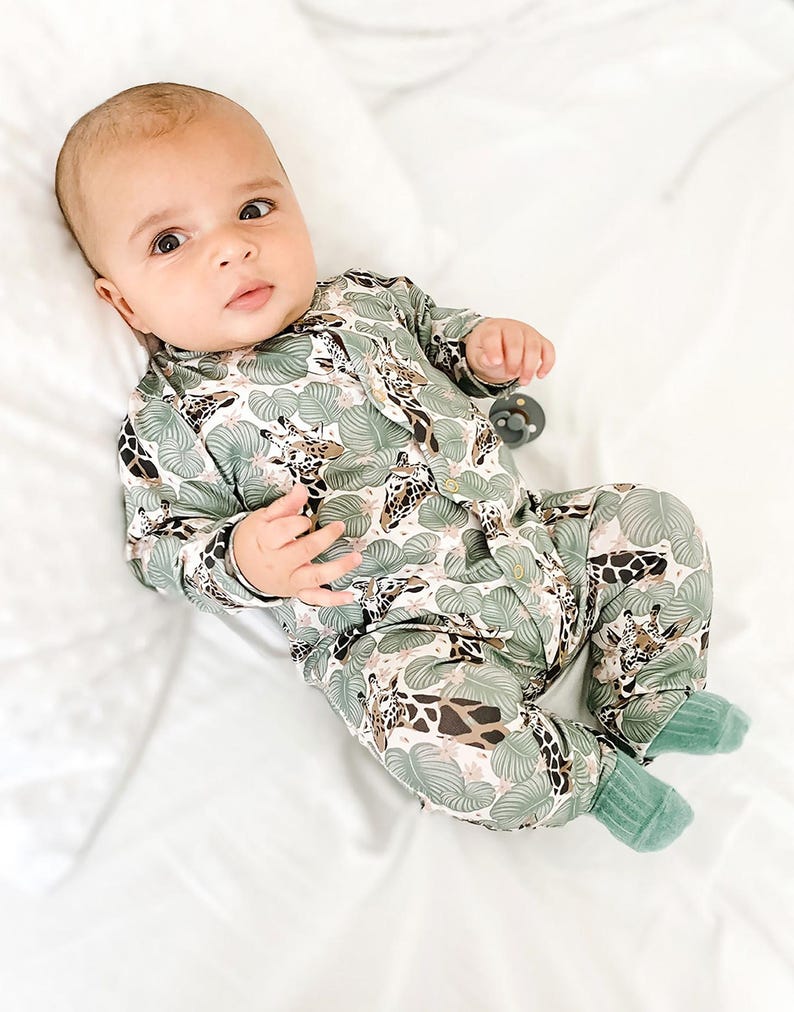 May include: A long-sleeved baby romper featuring a repeating pattern of giraffes and large green tropical leaves on a light background. The garment includes a front button closure and solid green ribbed cuffs at the ankles for a secure fit.
