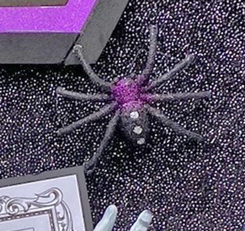 Puede incluir: Una ara&ntilde;a negra con un cuerpo morado y blanco y puntos blancos en sus patas. La ara&ntilde;a est&aacute; sobre un fondo negro con una textura brillante.