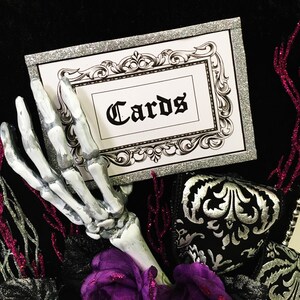 Puede incluir: Una caja de tarjetas de purpurina plateada con una tarjeta blanca que dice "Cards" en letra negra. Una mano esquel&eacute;tica sostiene la caja de tarjetas. La caja est&aacute; decorada con tela negra y plateada y una flor morada.