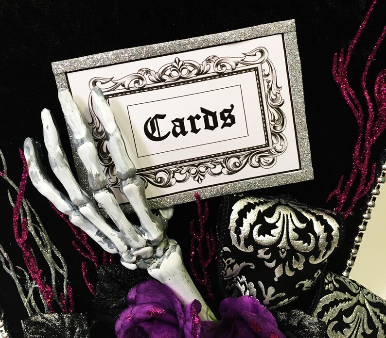 Puede incluir: Una caja de tarjetas de purpurina plateada con una tarjeta blanca que dice "Cards" en letra negra. Una mano esquel&eacute;tica sostiene la caja de tarjetas. La caja est&aacute; decorada con tela negra y plateada y una flor morada.