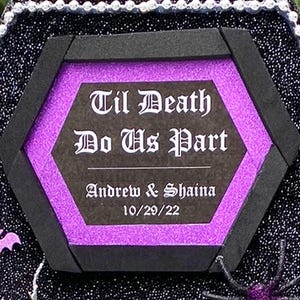 Puede incluir: Un letrero hexagonal negro con un fondo de purpurina morado y una inscripci&oacute;n blanca que dice "Til Death Do Us Part" con los nombres "Andrew & Shaina" y la fecha "10/29/22" debajo.