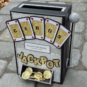May include: A black and silver decorative box designed to resemble a slot machine. The box features the word "JACKPOT" in gold lettering, playing card accents, and a lever with a glitter ball. Gold coins are visible in the payout tray.