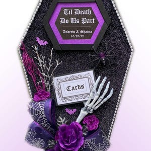 Puede incluir: Una caja de tarjetas negra en forma de ata&uacute;d con un letrero hexagonal morado y negro que dice "Til Death Do Us Part" con los nombres Andrew & Shaina y la fecha 10/29/22. La caja est&aacute; decorada con un lazo morado, telara&ntilde;as, murci&eacute;lagos y una mano de esqueleto. El letrero "Cards" est&aacute; en un marco blanco.