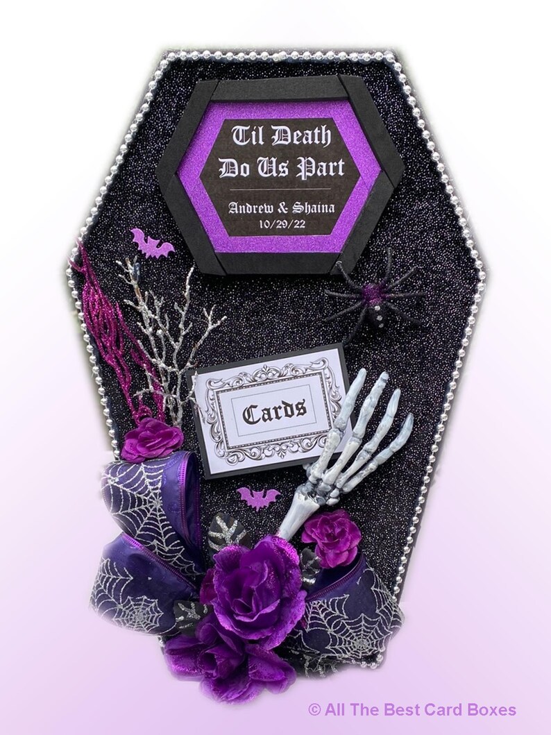 Puede incluir: Una caja de tarjetas negra en forma de ata&uacute;d con un letrero hexagonal morado y negro que dice "Til Death Do Us Part" con los nombres Andrew & Shaina y la fecha 10/29/22. La caja est&aacute; decorada con un lazo morado, telara&ntilde;as, murci&eacute;lagos y una mano de esqueleto. El letrero "Cards" est&aacute; en un marco blanco.