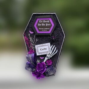 Puede incluir: Una caja de tarjetas negra en forma de ata&uacute;d con un lazo morado y negro, una mano de esqueleto, una ara&ntilde;a y un letrero que dice "Til Death Do Us Part" con los nombres Andrew & Shaina y la fecha 10/29/22. El letrero tambi&eacute;n dice "Cards".
