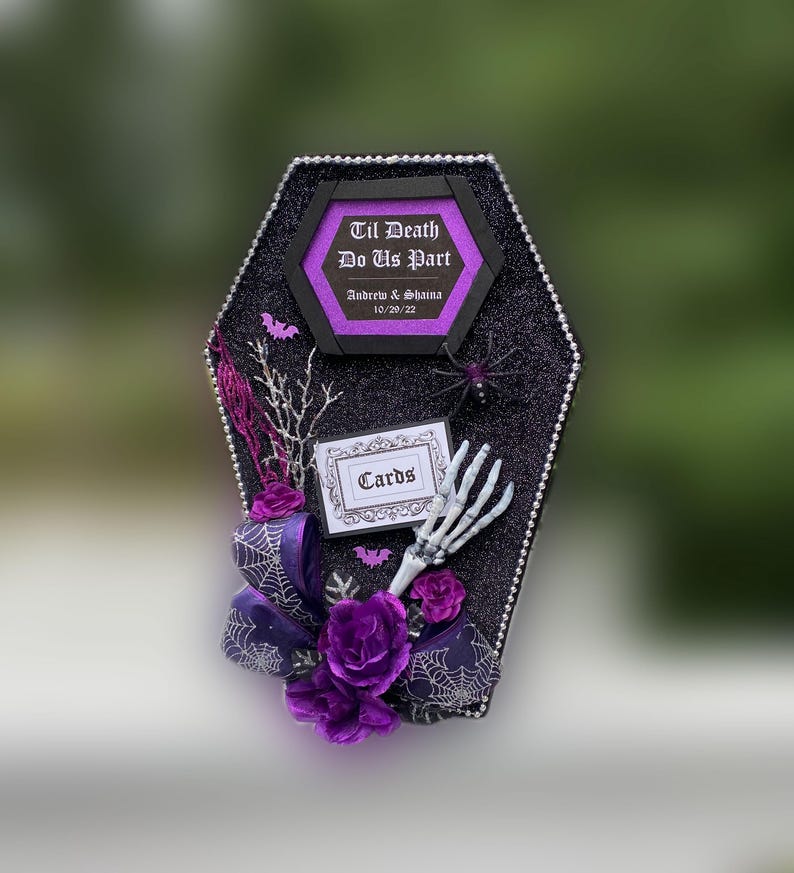 Puede incluir: Una caja de tarjetas negra en forma de ata&uacute;d con un lazo morado y negro, una mano de esqueleto, una ara&ntilde;a y un letrero que dice "Til Death Do Us Part" con los nombres Andrew & Shaina y la fecha 10/29/22. El letrero tambi&eacute;n dice "Cards".