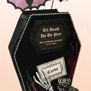 Puede incluir: Caja de tarjetas negra en forma de ata&uacute;d con un borde de cuentas plateadas. La caja tiene un letrero que dice "Til Death Do Us Part" con los nombres Ally y Douglas Reynolds y la fecha 10/31/2021. Una mano de esqueleto sale de la caja con un letrero que dice "Cards".