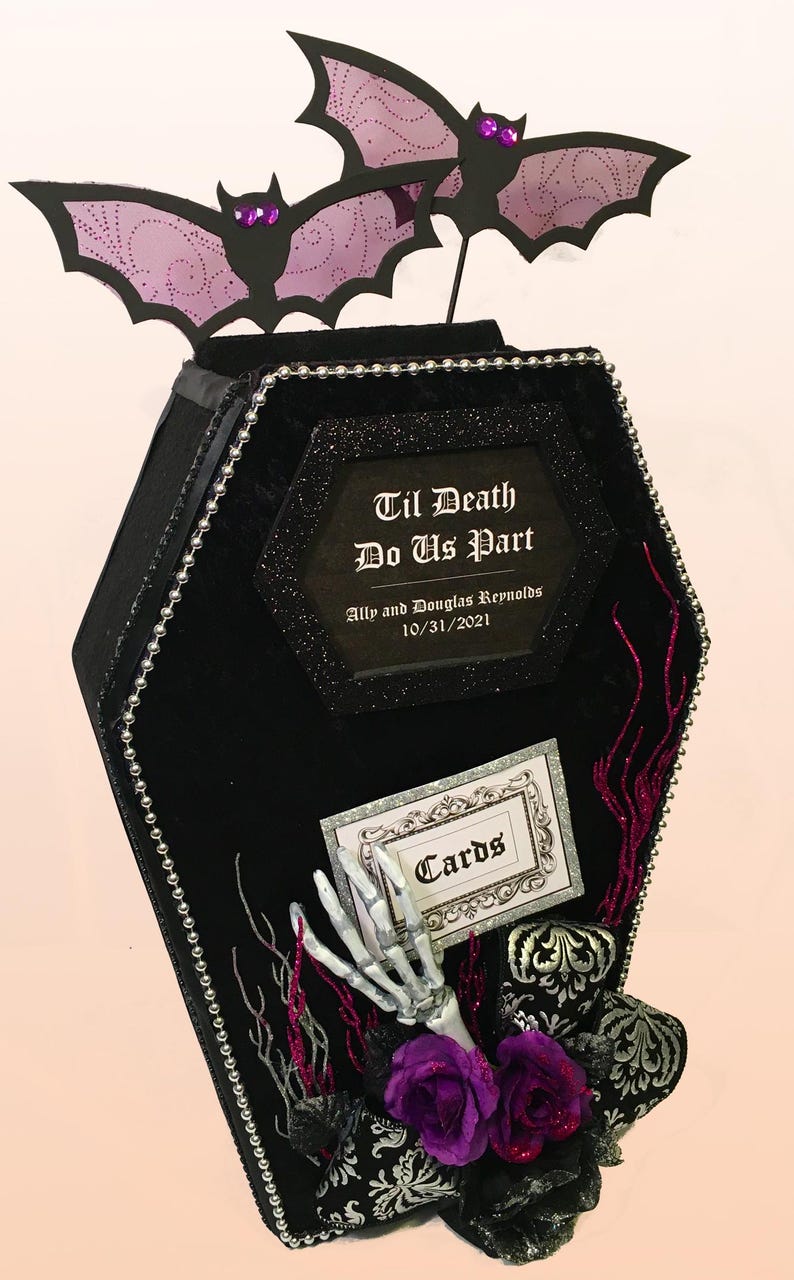 Puede incluir: Caja de tarjetas negra en forma de ata&uacute;d con un borde de cuentas plateadas. La caja tiene un letrero que dice "Til Death Do Us Part" con los nombres Ally y Douglas Reynolds y la fecha 10/31/2021. Una mano de esqueleto sale de la caja con un letrero que dice "Cards".