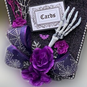 Puede incluir: Una caja negra con forma de ata&uacute;d con un lazo morado, una mano de esqueleto y flores moradas. La caja tiene un letrero que dice "Cartas".