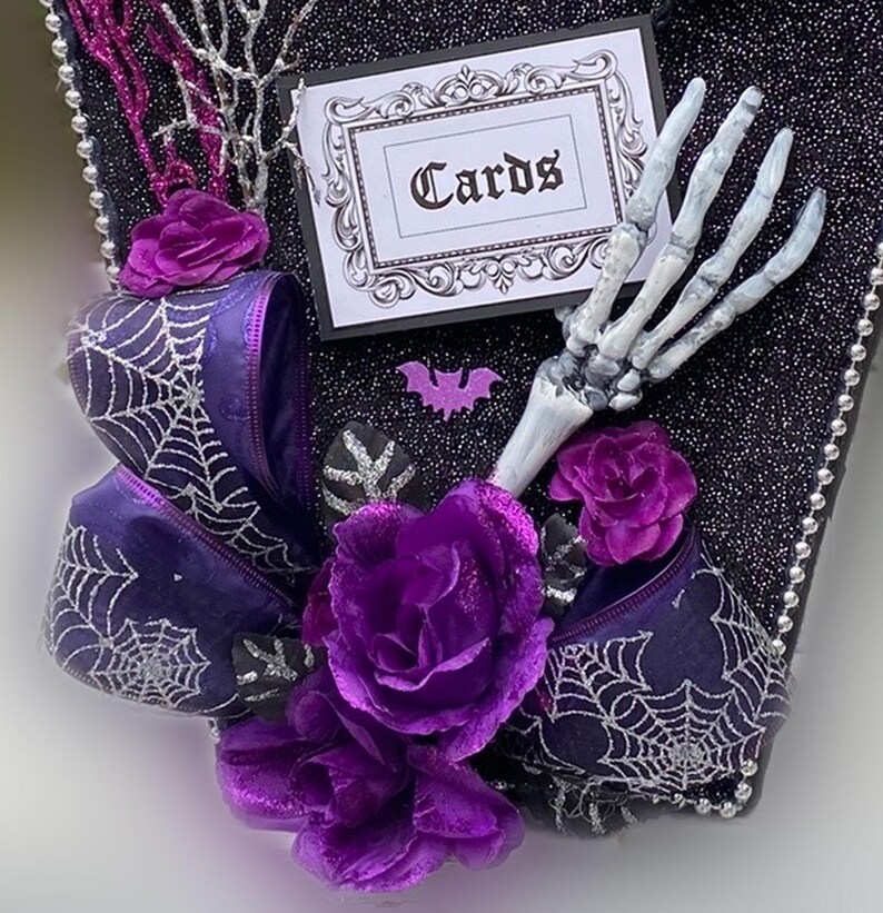 Puede incluir: Una caja negra con forma de ata&uacute;d con un lazo morado, una mano de esqueleto y flores moradas. La caja tiene un letrero que dice "Cartas".