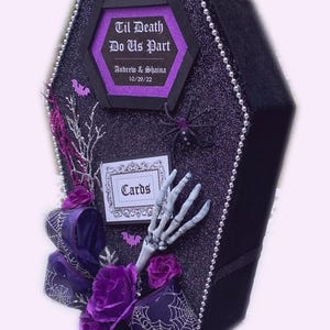 Puede incluir: Una caja de tarjetas negra en forma de ata&uacute;d con un acabado de purpurina morada. La caja est&aacute; decorada con una telara&ntilde;a blanca, una mano de esqueleto y un lazo morado. La caja tiene un letrero que dice "Til Death Do Us Part" con los nombres "Andrew & Shaina" y la fecha "10/20/22".