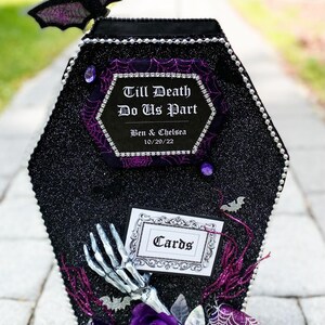 Puede incluir: Una caja de tarjetas negra en forma de ata&uacute;d con un acabado brillante y detalles morados. La caja tiene un letrero que dice "Till Death Do Us Part" con los nombres Ben & Chelsea y la fecha 10/29/22. La caja est&aacute; decorada con murci&eacute;lagos, telara&ntilde;as y flores.